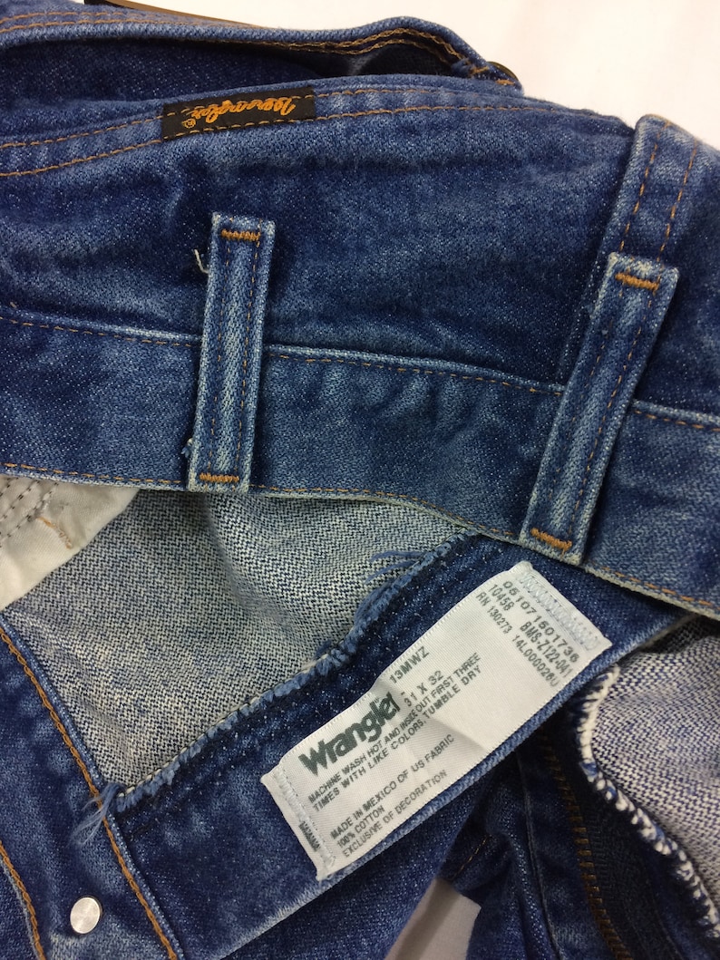 May include: A blue denim Wrangler jeans label with white text. The label reads "Machine wash hot and inside out first three times with like colors. Tumble dry. Made in Mexico of US fabric. 100% cotton. Exclusive of decoration."