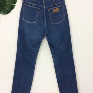 May include: Dark blue denim jeans with a classic five-pocket design. The jeans feature the Wrangler logo on a brown leather patch and on the back pockets. The jeans are hanging on a wooden hanger.