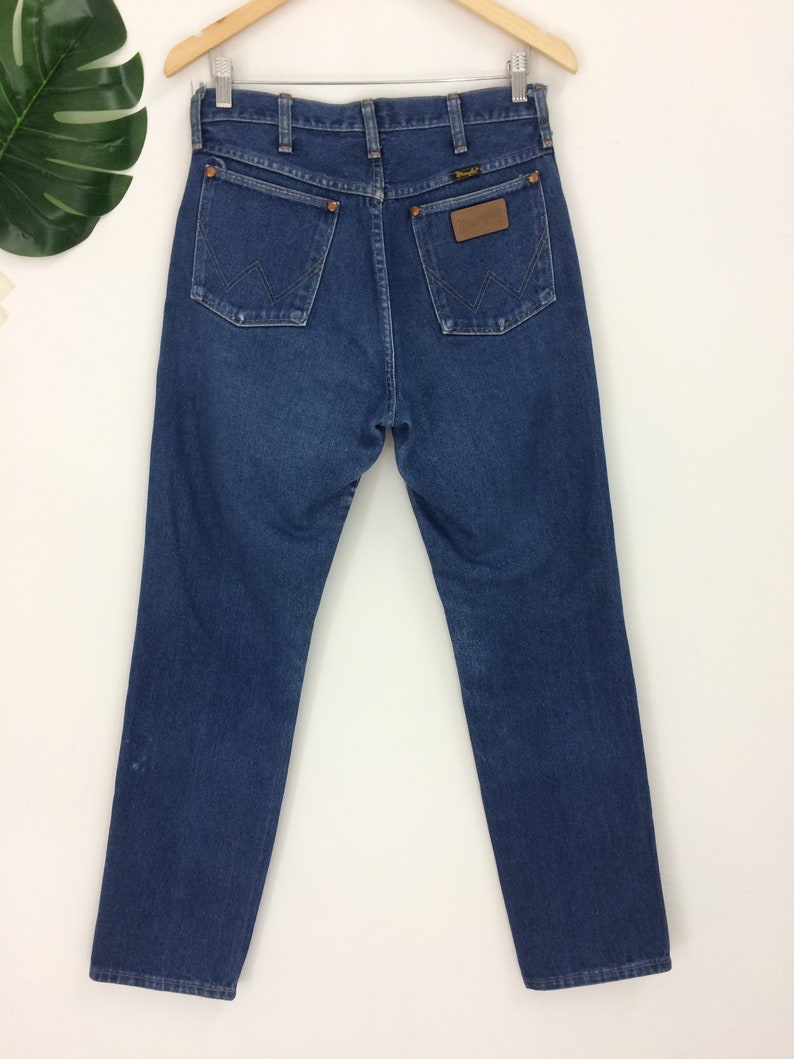 May include: Dark blue denim jeans with a classic five-pocket design. The jeans feature the Wrangler logo on a brown leather patch and on the back pockets. The jeans are hanging on a wooden hanger.