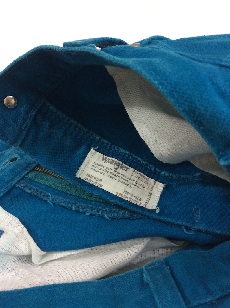 May include: A teal blue pair of Wrangler jeans with a white care label that reads "Machine wash with like colors, only non-chlorine bleach when needed, tumble dry, remove promptly. Made in USA 100% Cotton. M486-018 0 84084 324023"