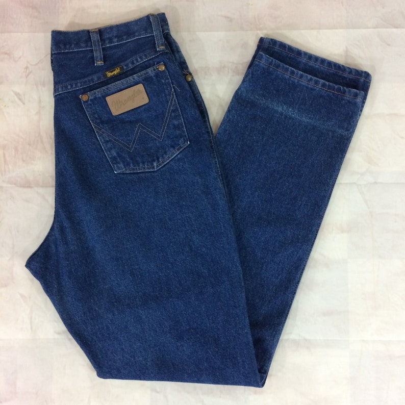 May include: Blue denim jeans with the Wrangler logo on the back pocket. The jeans have a classic five-pocket design, a button and zipper closure, and a straight leg fit. The jeans are a medium wash and appear to be in good condition.