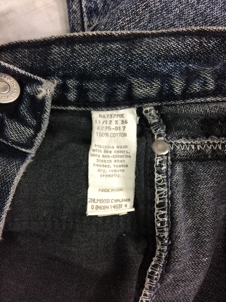 May include: Close-up of dark wash denim jeans. The tag reads "NA732MK 11/12 X 36 A275-017 100% COTTON". The jeans have a button closure and a stitched seam. The tag also includes washing instructions and "MADE IN USA".