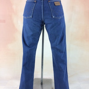 May include: Back view of blue denim jeans with the Wrangler logo on the back pocket. The jeans have a classic five-pocket design and a straight leg fit. The jeans are displayed on a metal stand.