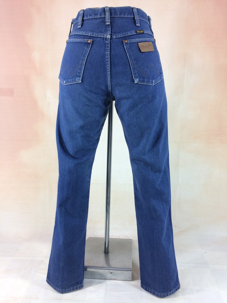 May include: Back view of blue denim jeans with the Wrangler logo on the back pocket. The jeans have a classic five-pocket design and a straight leg fit. The jeans are displayed on a metal stand.