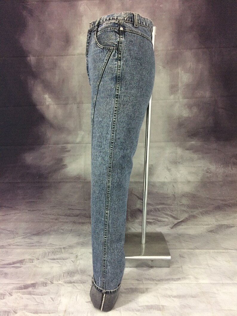 May include: Acid wash denim jeans with a unique diagonal seam detail on the front. The jeans have a classic five-pocket design and a straight leg fit. The hem is cuffed with a dark gray fabric.