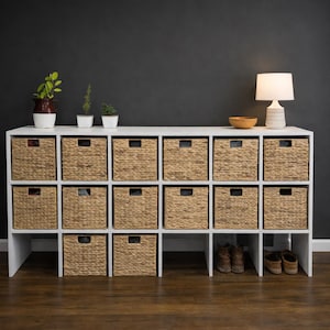 Puede incluir: Un mueble de almacenamiento blanco con cestas tejidas y una lámpara. La unidad tiene 15 cestas tejidas con asas cuadradas. La parte superior de la unidad tiene una lámpara, un cuenco de madera y pequeñas plantas en macetas. La unidad está contra una pared gris oscuro.