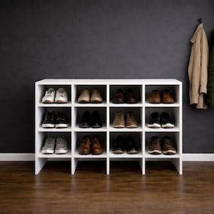 May include: A white shoe rack with multiple compartments, filled with various pairs of shoes. The shoes are in different styles and colors. A tan coat and a green jacket hang on the wall behind the rack.