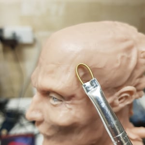 Könnte beinhalten: Nahaufnahme eines modellierten menschlichen Kopfes mit einer goldenen Drahtschlaufe, die an der Stirn befestigt ist. Ein Pinsel wird verwendet, um die Drahtschlaufe anzubringen.