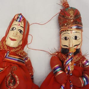 Puede incluir: Dos marionetas indias tradicionales. Las marionetas tienen caras de madera pintadas y están vestidas con tela roja con adornos dorados y arcoíris. Una marioneta lleva un tocado con lentejuelas.