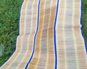 May include: A woven mat made of natural materials with a blue trim. The mat is folded in thirds and has a striped pattern in shades of beige, yellow, and green.