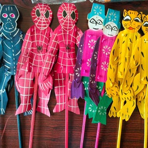 Op de afbeelding: Een set van acht kleurrijke houten poppen. De poppen zijn handgeschilderd en hebben verschillende ontwerpen, waaronder skeletten, spinnen, tijgers en bloemen. De poppen zijn bevestigd aan stokken.