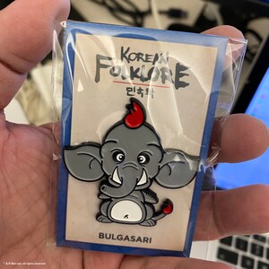 Puede incluir: Un pin de esmalte gris y rojo que representa un elefante de dibujos animados con una cresta roja, colmillos y cola. El pin está etiquetado como "BULGASARI" y "KOREAN FOLKLORE" en texto negro. El texto coreano "민속화" también está incluido.