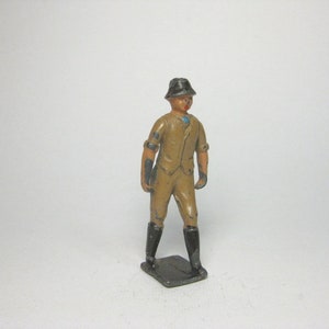 Puede incluir: Una figura de juguete de plomo vintage de una persona que lleva un atuendo marrón y negro, un sombrero negro y botas negras. La figura está de pie sobre una base negra.
