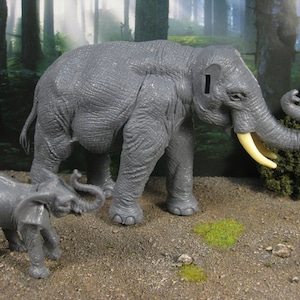 May include: A grey toy elephant with a smaller grey toy elephant calf standing next to it. The larger elephant has a long trunk and large ears. The smaller elephant has a shorter trunk and smaller ears. Both elephants are standing on a brown surface with green grass.