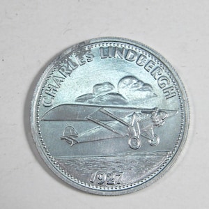 Puede incluir: Una moneda de plata con la inscripción "CHARLES LINDBERGH" y el año "1927". La moneda representa un avión volando sobre agua y nubes. La moneda es redonda con un borde texturizado y el diseño es muy detallado.