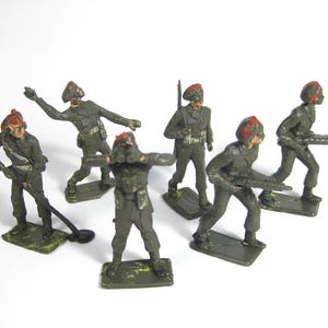 May include: Six green plastic toy soldiers wearing red and white hats. The soldiers are holding rifles and are in various poses.