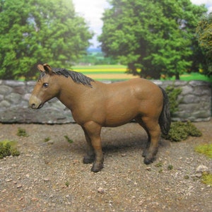 Pode incluir: Uma figura de cavalo marrom com crina e cauda pretas. A figura está em pé em uma superfície de cascalho com uma parede de pedra no fundo.