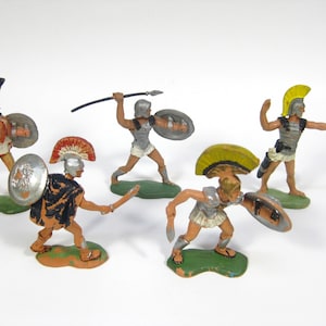 May include: Five plastic toy figurines of ancient Greek warriors. The figures are painted in shades of brown, yellow, red, and silver. Each figure is holding a weapon, such as a sword, spear, or bow and arrow.