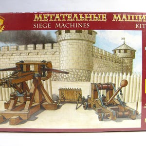 Può includere: Scatola di un modellino intitolata "SIEGE MACHINES" e "KIT No.1". L'immagine sulla scatola raffigura una scena di castello medievale con due macchine d'assedio in legno. La scatola è rossa con scritte dorate e include il testo "MODEL KIT". Scala 1/72.