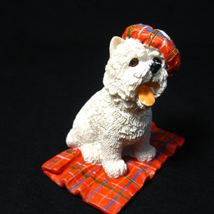 Puede incluir: Una figurita blanca de un perro con un sombrero de cuadros rojos, azules y verdes. El perro está sentado sobre una manta de cuadros rojos, azules y verdes.