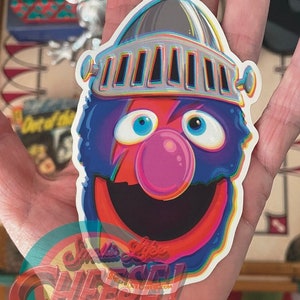 May include: A blue and red character with a large smile and a silver helmet with a rainbow effect. The character has a pink nose and large blue eyes. The sticker has a rainbow border and the text "Smells Like Cheese!" at the bottom.
