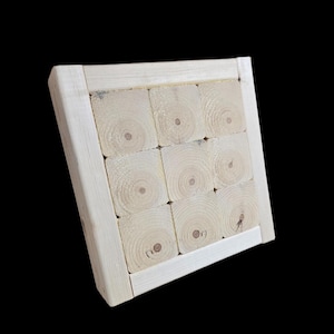 May include: A light-colored wooden square, constructed from nine square blocks of wood, each displaying circular growth rings. The blocks are framed by a lighter-colored wooden border. The background is solid black.