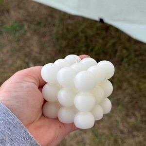 May include: A white, cube-shaped candle composed of numerous spherical wax bubbles. The candle is held in a hand, with a blurred outdoor background of grass and a white canopy.