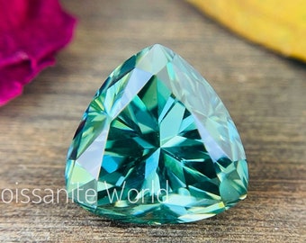 May include: A triangular cut green moissanite gemstone with a faceted design. The gemstone is set against a wooden background with a pink flower petal and a yellow leaf.