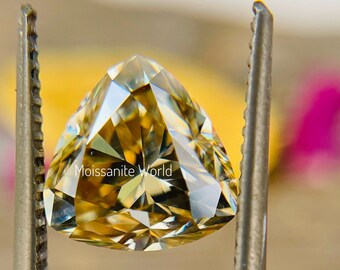 Puede incluir: Una piedra preciosa de moissanita de corte triangular, de color dorado, sostenida por unas pinzas. La piedra preciosa tiene un corte brillante y facetado y un tono dorado cálido. El texto "Moissanite World" es visible en la imagen.