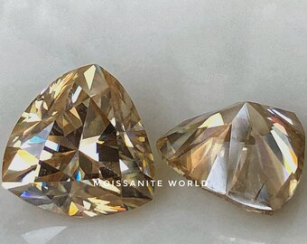 Puede incluir: Dos piedras preciosas talladas en forma triangular, una más grande que la otra, de color champán y un brillo brillante. Las piedras preciosas están sobre una superficie blanca. "Moissanite World" es visible en la imagen.
