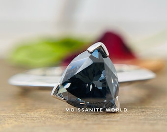 Pode incluir: Um anel de prata com uma gema triangular de cor cinza escura. O anel está colocado sobre uma superfície de madeira. O texto "Moissanite World" é visível abaixo do anel.