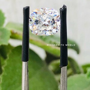 May include: A clear, cushion-cut moissanite gemstone held by tweezers against a green, blurred background. The text "Moissanite World" is visible below the gemstone.