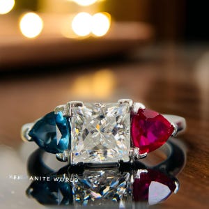 May include: A silver ring with a large square-shaped diamond in the center, flanked by a blue sapphire on the left and a red ruby on the right.