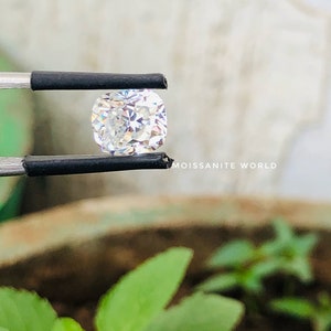 May include: A clear, square-shaped, faceted gemstone held by tweezers. The gemstone is reflecting light, creating a rainbow effect. The background is blurred and shows green foliage.