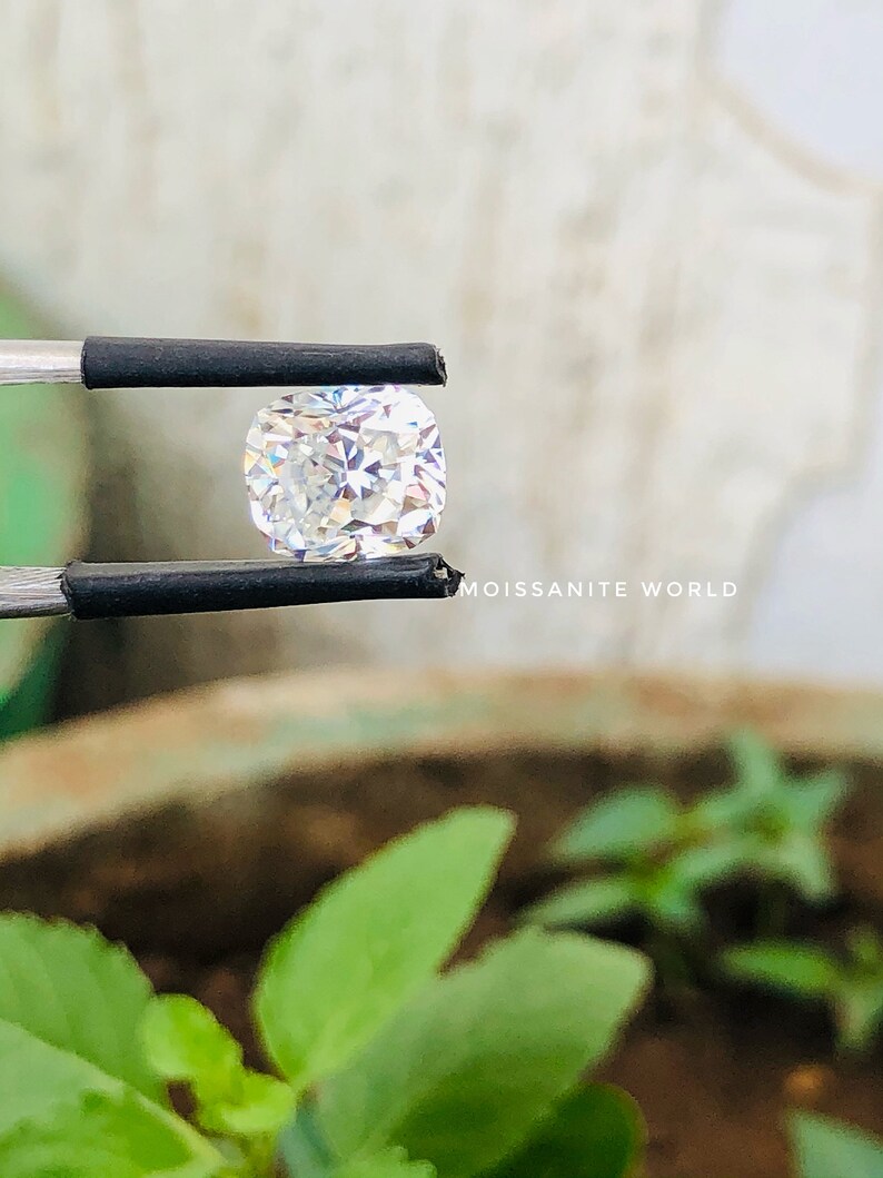 May include: A clear, square-shaped, faceted gemstone held by tweezers. The gemstone is reflecting light, creating a rainbow effect. The background is blurred and shows green foliage.