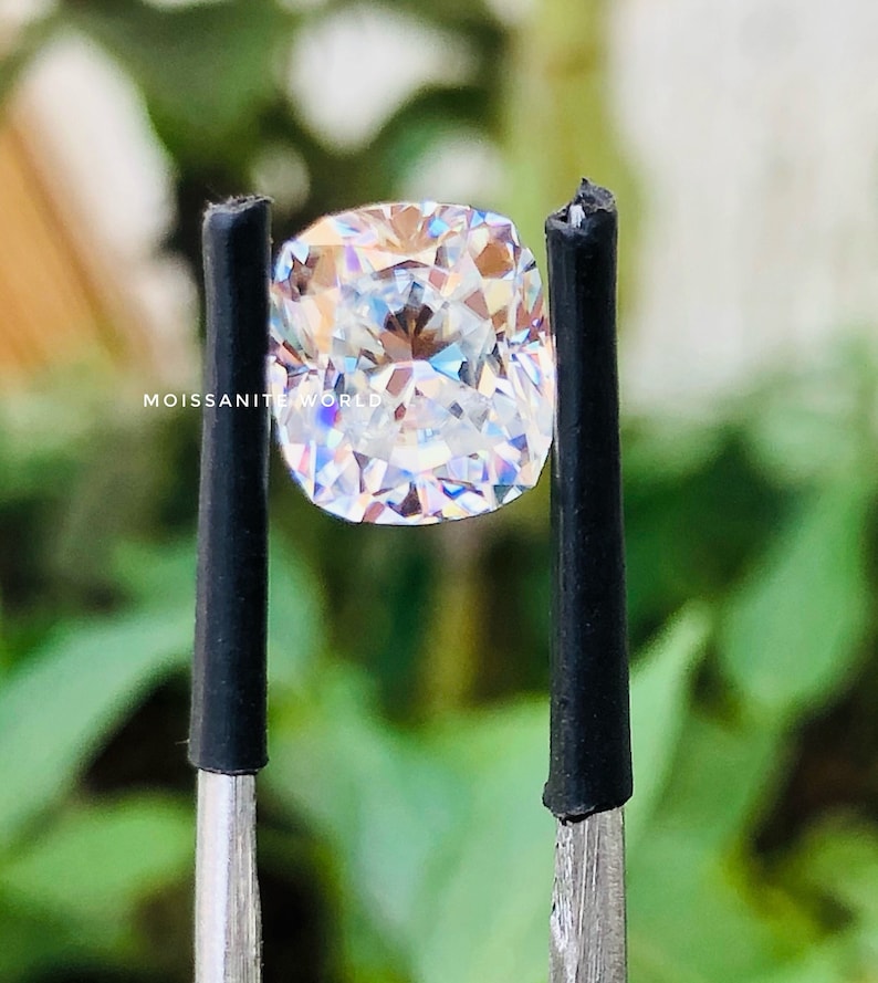 May include: A clear, square-shaped, cushion cut gemstone with a rainbow of colors reflecting off its facets. The gemstone is held by two black tweezers.