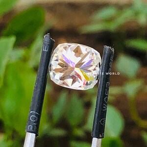 May include: A clear, faceted, square-shaped gemstone with rainbow-like reflections. The gemstone is held by a pair of black tweezers. The text "Moissanite World" is visible in the background.