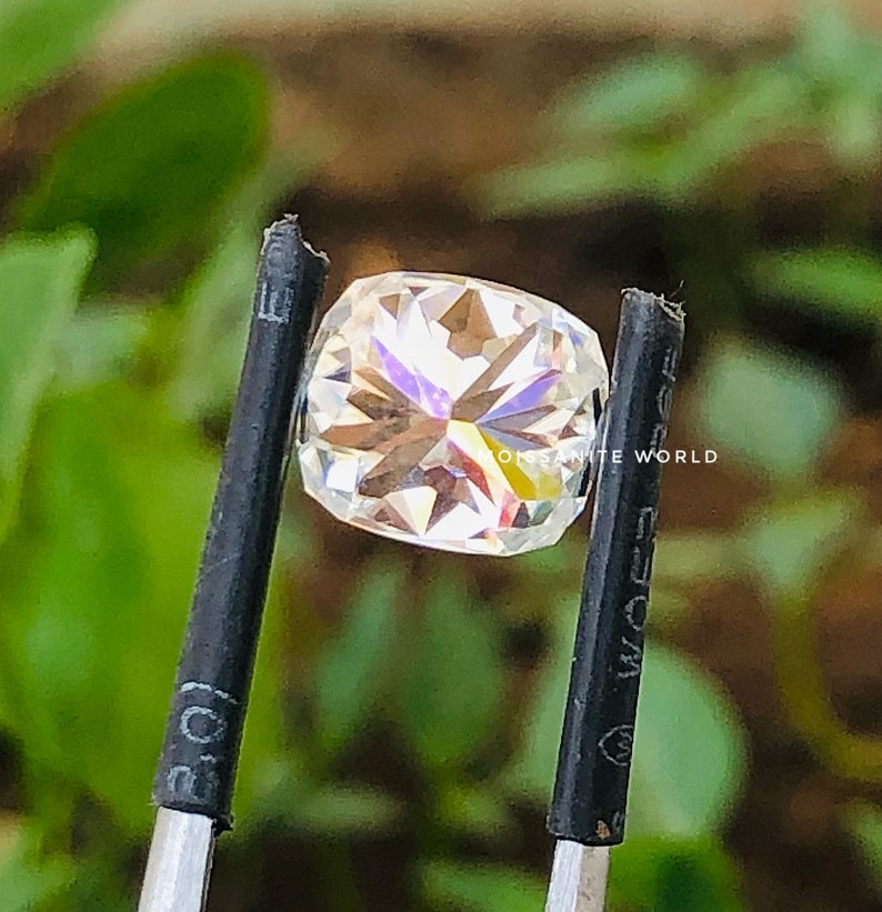 May include: A clear, faceted, square-shaped gemstone with rainbow-like reflections. The gemstone is held by a pair of black tweezers. The text "Moissanite World" is visible in the background.