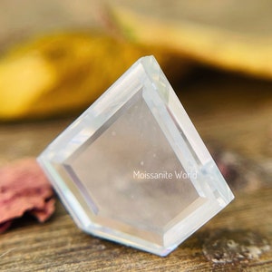 K&ouml;nnte beinhalten: Ein klarer, facettierter, diamantf&ouml;rmiger Edelstein mit einer abgeschr&auml;gten Kante. Der Edelstein liegt auf einer Holzoberfl&auml;che. Der Text "Moissanite World" ist in der Mitte des Edelsteins sichtbar.