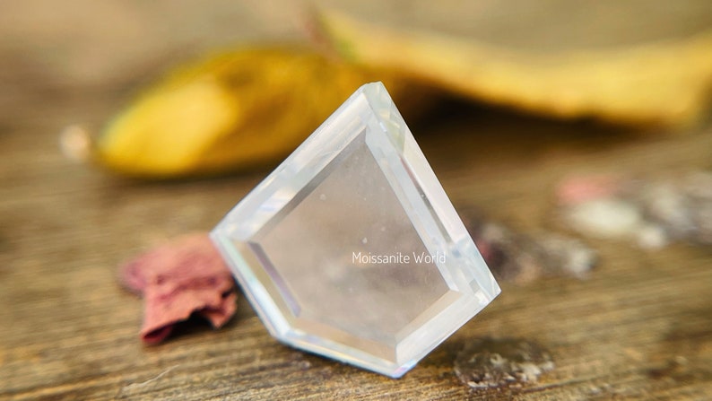K&ouml;nnte beinhalten: Ein klarer, facettierter, diamantf&ouml;rmiger Edelstein mit einer abgeschr&auml;gten Kante. Der Edelstein liegt auf einer Holzoberfl&auml;che. Der Text "Moissanite World" ist in der Mitte des Edelsteins sichtbar.