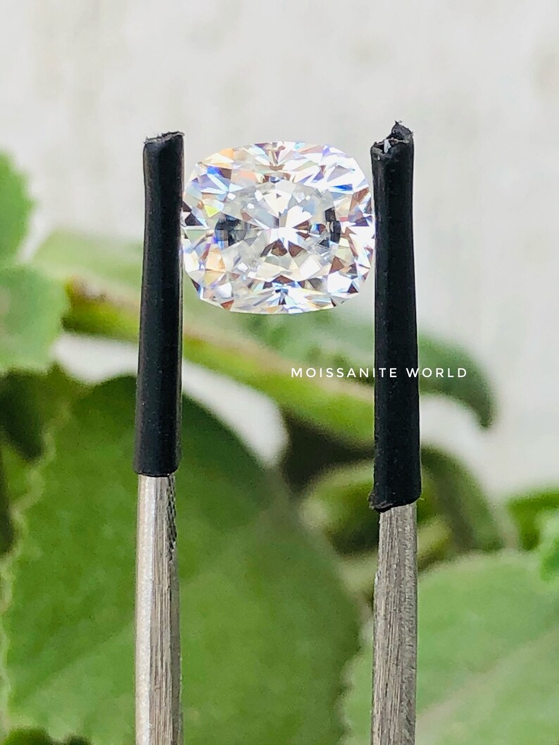 May include: A clear, cushion-cut moissanite gemstone held by tweezers against a green, blurred background. The text "Moissanite World" is visible below the gemstone.