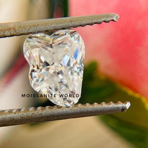 May include: A clear heart-shaped moissanite gemstone held by a pair of tweezers. The gemstone is faceted and reflects light. The background is blurred and out of focus.