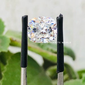 May include: A clear, cushion-cut moissanite gemstone is held by a pair of tweezers. The gemstone is reflecting light, showing its brilliance and facets. The background is blurred, with green foliage visible.