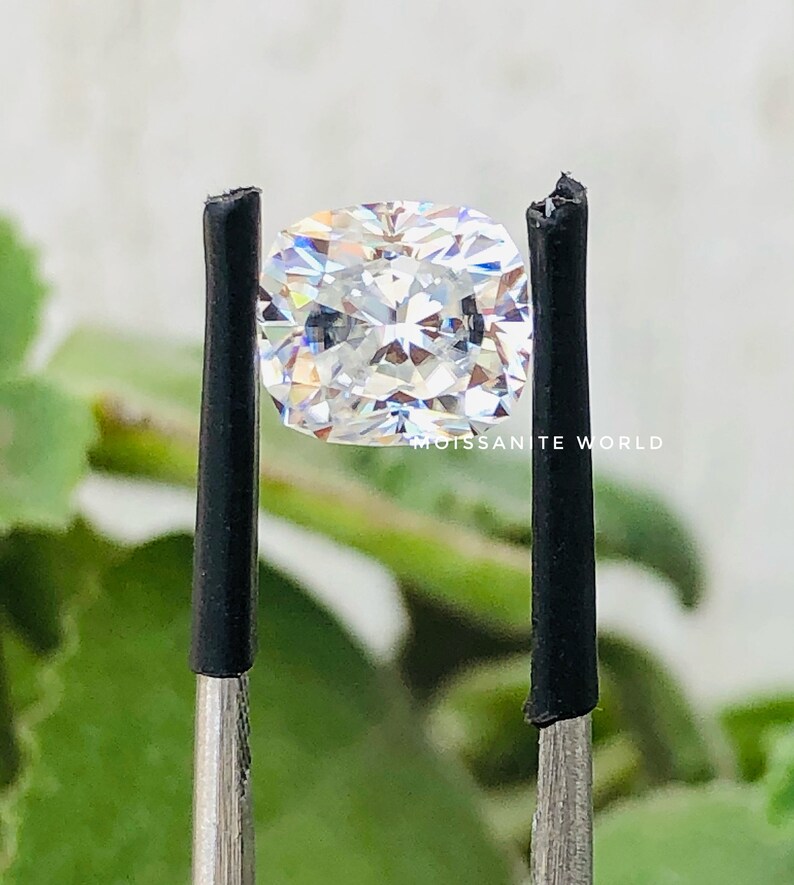 May include: A clear, cushion-cut moissanite gemstone is held by a pair of tweezers. The gemstone is reflecting light, showing its brilliance and facets. The background is blurred, with green foliage visible.