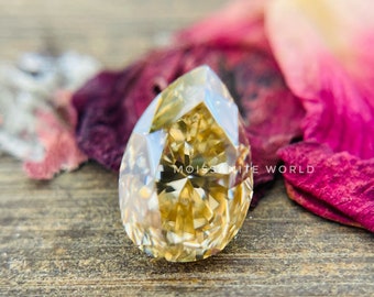 May include: A pear-shaped, golden moissanite gemstone with a brilliant cut, resting on a pink flower petal.