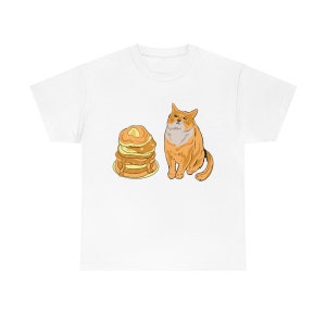 Pode incluir: Uma camiseta branca com uma imagem de desenho animado de um gato olhando para uma pilha de panquecas com xarope.