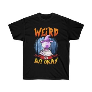 Puede incluir: Camiseta negra con un gráfico de una bruja sosteniendo una bola de cristal y el texto "Weird but okay" con la palabra "hex" debajo de la bola de cristal.