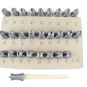 May include: A set of 28 metal letter stamps for stamping leather or metal. The stamps are arranged in alphabetical order on a beige leatherette backing. A wooden handle with a metal stamp head is also included.