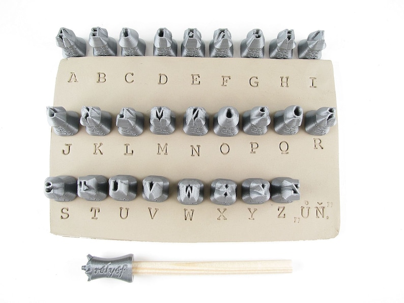 May include: A set of 28 metal letter stamps for stamping leather or metal. The stamps are arranged in alphabetical order on a beige leatherette backing. A wooden handle with a metal stamp head is also included.