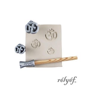 Könnte beinhalten: Ein Set aus Om-Symbolstempeln und einem Holzgriff mit dem Markennamen "rélyéf". Die Stempel sind grau und der Griff ist hellbraun. Das Om-Symbol ist auf einem beigen Block aufgedruckt.